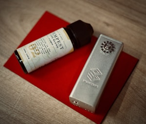 A cylindrical vape juice bottle with a light-colored label and a black cap is placed next to a metallic vape mod. Both items are resting on a red rectangular surface that contrasts with the wooden tabletop underneath.