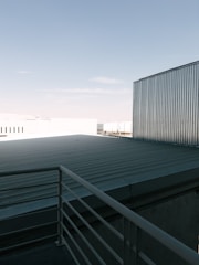 A rooftop with a metal railing and corrugated metal sheets under a clear blue sky. The scene includes industrial-looking buildings with metallic textures.