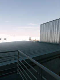 A rooftop with a metal railing and corrugated metal sheets under a clear blue sky. The scene includes industrial-looking buildings with metallic textures.