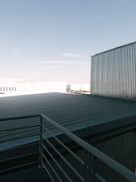 A rooftop with a metal railing and corrugated metal sheets under a clear blue sky. The scene includes industrial-looking buildings with metallic textures.