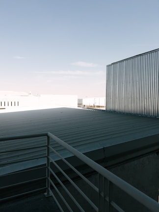 A rooftop with a metal railing and corrugated metal sheets under a clear blue sky. The scene includes industrial-looking buildings with metallic textures.
