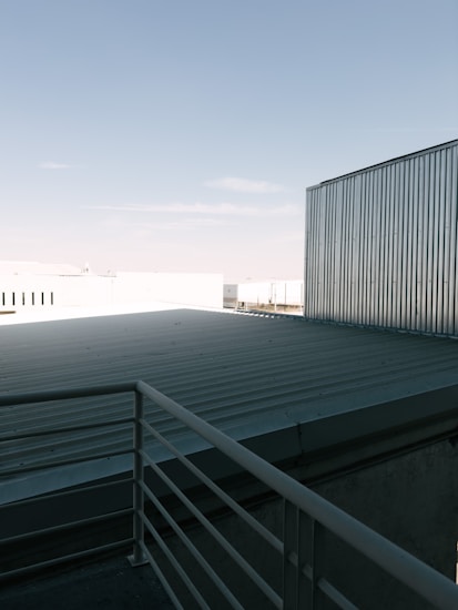 A rooftop with a metal railing and corrugated metal sheets under a clear blue sky. The scene includes industrial-looking buildings with metallic textures.