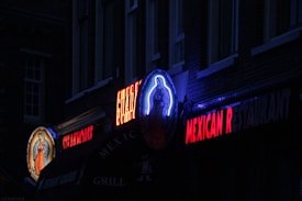 A dimly lit street scene featuring neon signs for a Mexican restaurant. A vivid religious icon is illuminated on the left, and vibrant red lettering spells out 'MEXICAN RESTAURANT'. The atmosphere appears dark with hints of blue and red lighting.