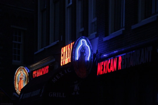 A dimly lit street scene featuring neon signs for a Mexican restaurant. A vivid religious icon is illuminated on the left, and vibrant red lettering spells out 'MEXICAN RESTAURANT'. The atmosphere appears dark with hints of blue and red lighting.