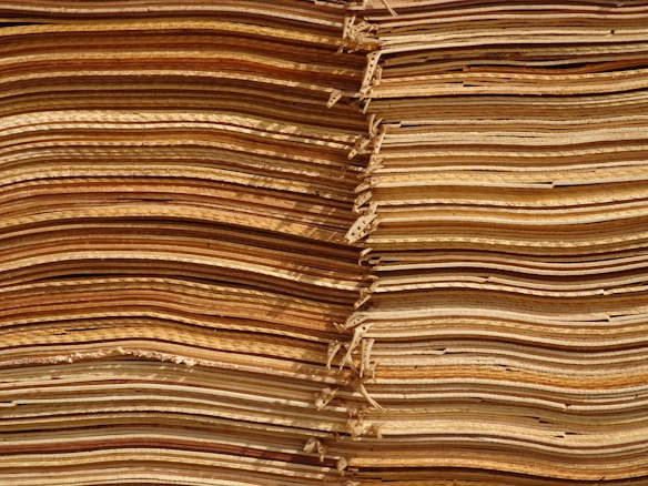 Stacks of thin, unevenly cut wooden sheets create a visually intricate and textured pattern. The sheets vary in color, ranging from light tan to deep brown, and appear to be layered in an organized manner.