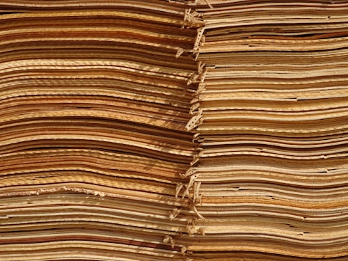 Stacks of thin, unevenly cut wooden sheets create a visually intricate and textured pattern. The sheets vary in color, ranging from light tan to deep brown, and appear to be layered in an organized manner.