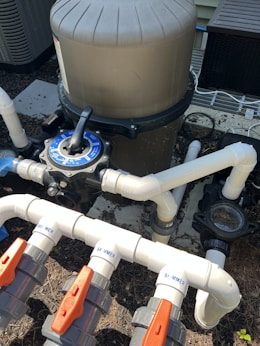 A complex system of pipes and valves is connected to a large, cylindrical filtration unit, likely part of a pool system. White PVC pipes with red and gray valve handles are labeled 'SKIMMER' and are arranged at different angles. The surrounding area includes mulch and parts of an air conditioning unit.