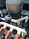 A complex system of pipes and valves is connected to a large, cylindrical filtration unit, likely part of a pool system. White PVC pipes with red and gray valve handles are labeled 'SKIMMER' and are arranged at different angles. The surrounding area includes mulch and parts of an air conditioning unit.