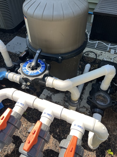 A complex system of pipes and valves is connected to a large, cylindrical filtration unit, likely part of a pool system. White PVC pipes with red and gray valve handles are labeled 'SKIMMER' and are arranged at different angles. The surrounding area includes mulch and parts of an air conditioning unit.