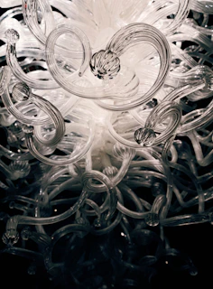 A sculptural installation featuring intertwined metal and glass elements reflecting soft gallery lighting.