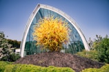 A vibrant glass sculpture featuring an explosion of yellow and orange swirls is prominently displayed in front of a modern, arched glass building. The structure behind is surrounded by lush greenery, suggesting a garden or botanical setting. The sky is clear and blue, adding to the bright, lively atmosphere.