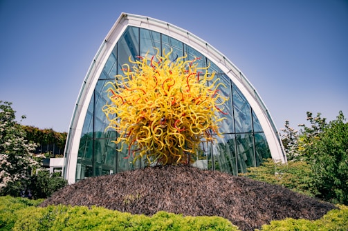 A vibrant glass sculpture featuring an explosion of yellow and orange swirls is prominently displayed in front of a modern, arched glass building. The structure behind is surrounded by lush greenery, suggesting a garden or botanical setting. The sky is clear and blue, adding to the bright, lively atmosphere.