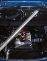 A close-up of a car engine under the hood, featuring a metallic support bar and visible wiring. The engine parts are detailed, with components like pipes and wires prominently shown.