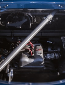 A close-up of a car engine under the hood, featuring a metallic support bar and visible wiring. The engine parts are detailed, with components like pipes and wires prominently shown.