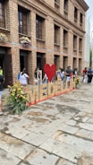 A large sign spelling 'Medellin' with a red heart is placed in front of a brick building. Several people are interacting with the area, and colorful flowers are arranged around the sign. The building features multiple windows with flower boxes.
