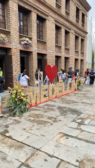 A large sign spelling 'Medellin' with a red heart is placed in front of a brick building. Several people are interacting with the area, and colorful flowers are arranged around the sign. The building features multiple windows with flower boxes.