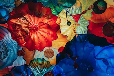 A vibrant display of colorful glass sculptures with intricate patterns and shapes, creating a mesmerizing kaleidoscope effect. The artwork features large, wavy forms in red, orange, blue, and green hues, along with various textures and designs.