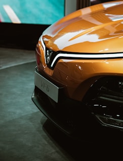 A sleek, modern car with a glossy orange exterior is prominently displayed. The front of the vehicle features a distinctive V-shaped logo and stylish headlights. The setting appears to be indoors, possibly at an auto show, and the car's polished surface reflects its surroundings.
