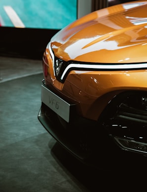 A sleek, modern car with a glossy orange exterior is prominently displayed. The front of the vehicle features a distinctive V-shaped logo and stylish headlights. The setting appears to be indoors, possibly at an auto show, and the car's polished surface reflects its surroundings.