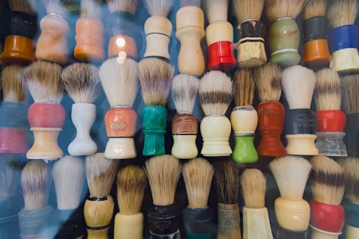 A collection of various shaving brushes neatly arranged in rows with different colored handles and bristle patterns. The handles are made of materials like plastic and wood, showing signs of wear and variety in design. Some brushes have a glossy finish while others appear matte.