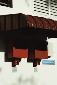 Two red electrical boxes are mounted on a white wall beneath a red corrugated iron awning. Below the boxes, small signs indicate electricity-related functions. A blue sign below reads 'PENCAWANG ELEKTRIK NO 11A'. The area is partly shadowed, emphasizing the geometric shapes.