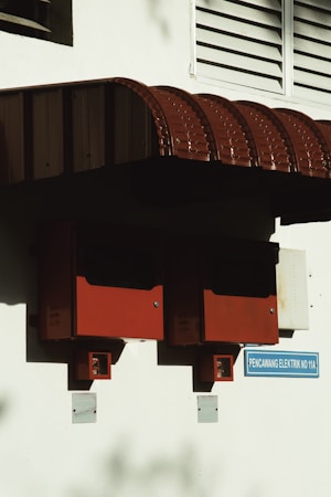 Two red electrical boxes are mounted on a white wall beneath a red corrugated iron awning. Below the boxes, small signs indicate electricity-related functions. A blue sign below reads 'PENCAWANG ELEKTRIK NO 11A'. The area is partly shadowed, emphasizing the geometric shapes.