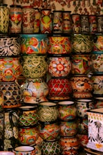 A collection of vibrant and intricately decorated ceramic pots and vases stacked on shelves. Each piece features unique patterns, including floral motifs, geometric designs, and colorful fish. The ceramics vary in size and shape, contributing to an eclectic and artistic display.