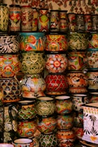 A collection of vibrant and intricately decorated ceramic pots and vases stacked on shelves. Each piece features unique patterns, including floral motifs, geometric designs, and colorful fish. The ceramics vary in size and shape, contributing to an eclectic and artistic display.