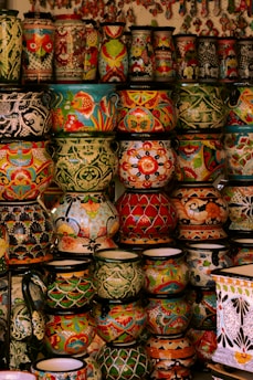 A collection of vibrant and intricately decorated ceramic pots and vases stacked on shelves. Each piece features unique patterns, including floral motifs, geometric designs, and colorful fish. The ceramics vary in size and shape, contributing to an eclectic and artistic display.