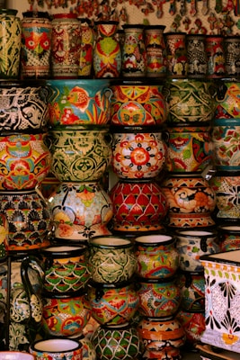 A collection of vibrant and intricately decorated ceramic pots and vases stacked on shelves. Each piece features unique patterns, including floral motifs, geometric designs, and colorful fish. The ceramics vary in size and shape, contributing to an eclectic and artistic display.