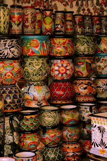 A collection of vibrant and intricately decorated ceramic pots and vases stacked on shelves. Each piece features unique patterns, including floral motifs, geometric designs, and colorful fish. The ceramics vary in size and shape, contributing to an eclectic and artistic display.