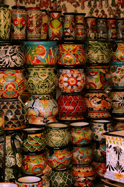A collection of vibrant and intricately decorated ceramic pots and vases stacked on shelves. Each piece features unique patterns, including floral motifs, geometric designs, and colorful fish. The ceramics vary in size and shape, contributing to an eclectic and artistic display.