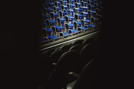 A view of numerous blue desks and chairs arranged in rows, likely in a classroom or exam hall setting. The image is taken from an obscured vantage point, with shadows enveloping the foreground.