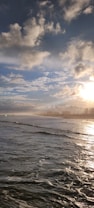 The image captures a serene beach scene with gentle waves and the warm glow of the setting sun reflecting on the water. Fluffy clouds fill the sky, and a distant shoreline with trees can be seen in the background.