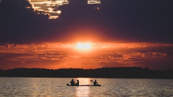 Silhouettes of two people kayaking on a calm lake during a dramatic sunset. The sky is filled with dark clouds, but the sun breaks through, creating a stunning contrast with warm orange and red hues. The water reflects these colors, adding to the serene yet powerful ambiance.