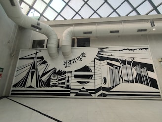 A large, monochromatic mural on an interior wall featuring abstract architectural designs and geometric shapes. The mural includes elements like stylized buildings and bridges, with bold black lines against a white background. Above the mural are visible air ducts leading to a glass-paned ceiling.