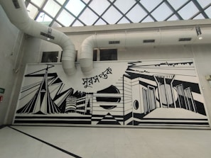 A large, monochromatic mural on an interior wall featuring abstract architectural designs and geometric shapes. The mural includes elements like stylized buildings and bridges, with bold black lines against a white background. Above the mural are visible air ducts leading to a glass-paned ceiling.