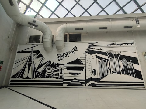A large, monochromatic mural on an interior wall featuring abstract architectural designs and geometric shapes. The mural includes elements like stylized buildings and bridges, with bold black lines against a white background. Above the mural are visible air ducts leading to a glass-paned ceiling.