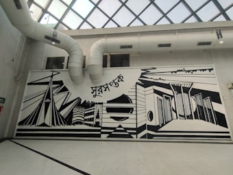 A large, monochromatic mural on an interior wall featuring abstract architectural designs and geometric shapes. The mural includes elements like stylized buildings and bridges, with bold black lines against a white background. Above the mural are visible air ducts leading to a glass-paned ceiling.