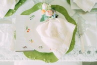 A beautifully set table with a white embroidered napkin atop a decorative plate featuring small fruit designs. The plate rests on a larger green ornamental charger. A floral and vegetable-themed napkin ring adds a touch of elegance. Silver cutlery, including a fork, knife, and a glimpse of a menu or card, complete the setting.