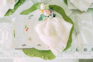 A beautifully set table with a white embroidered napkin atop a decorative plate featuring small fruit designs. The plate rests on a larger green ornamental charger. A floral and vegetable-themed napkin ring adds a touch of elegance. Silver cutlery, including a fork, knife, and a glimpse of a menu or card, complete the setting.