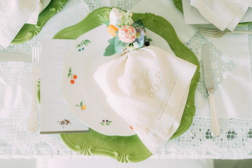A beautifully set table with a white embroidered napkin atop a decorative plate featuring small fruit designs. The plate rests on a larger green ornamental charger. A floral and vegetable-themed napkin ring adds a touch of elegance. Silver cutlery, including a fork, knife, and a glimpse of a menu or card, complete the setting.