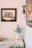 A decorative corner featuring a floral painting on the wall, a vase with flowers on a patterned tablecloth, and a shelf with a decorative mushroom ornament. The scene includes glassware on the table and a framed artwork with a fruit motif in the background.