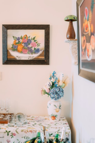 A decorative corner featuring a floral painting on the wall, a vase with flowers on a patterned tablecloth, and a shelf with a decorative mushroom ornament. The scene includes glassware on the table and a framed artwork with a fruit motif in the background.