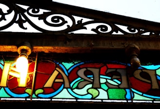 A vintage stained glass window with an ornate metal grille above it. The glass features vibrant colors including red, blue, green, and a light bulb lit on the left side, casting a warm glow. The metal grille has intricate, swirling patterns and a slightly weathered look.