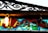 A vintage stained glass window with an ornate metal grille above it. The glass features vibrant colors including red, blue, green, and a light bulb lit on the left side, casting a warm glow. The metal grille has intricate, swirling patterns and a slightly weathered look.