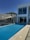 A luxurious modern house with a sleek infinity pool extending outward. Large glass windows dominate the facade, reflecting the clear blue sky. Minimalist design elements, including a shaded seating area and a simple railing, complement the clean architectural lines. Trees are visible just beyond the house, suggesting a serene surrounding.