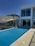 A luxurious modern house with a sleek infinity pool extending outward. Large glass windows dominate the facade, reflecting the clear blue sky. Minimalist design elements, including a shaded seating area and a simple railing, complement the clean architectural lines. Trees are visible just beyond the house, suggesting a serene surrounding.