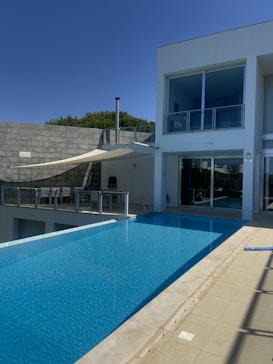 A luxurious modern house with a sleek infinity pool extending outward. Large glass windows dominate the facade, reflecting the clear blue sky. Minimalist design elements, including a shaded seating area and a simple railing, complement the clean architectural lines. Trees are visible just beyond the house, suggesting a serene surrounding.