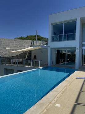A luxurious modern house with a sleek infinity pool extending outward. Large glass windows dominate the facade, reflecting the clear blue sky. Minimalist design elements, including a shaded seating area and a simple railing, complement the clean architectural lines. Trees are visible just beyond the house, suggesting a serene surrounding.